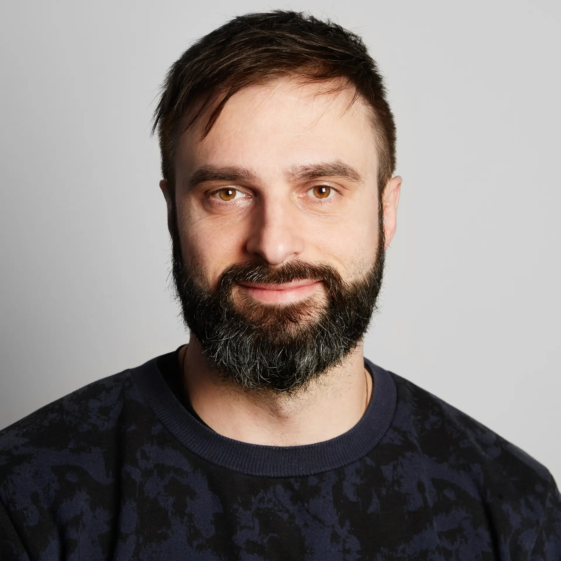 Foto de perfil de hombre de ~30 años, con barba cuidada, camisa oscura, fondo neutro.