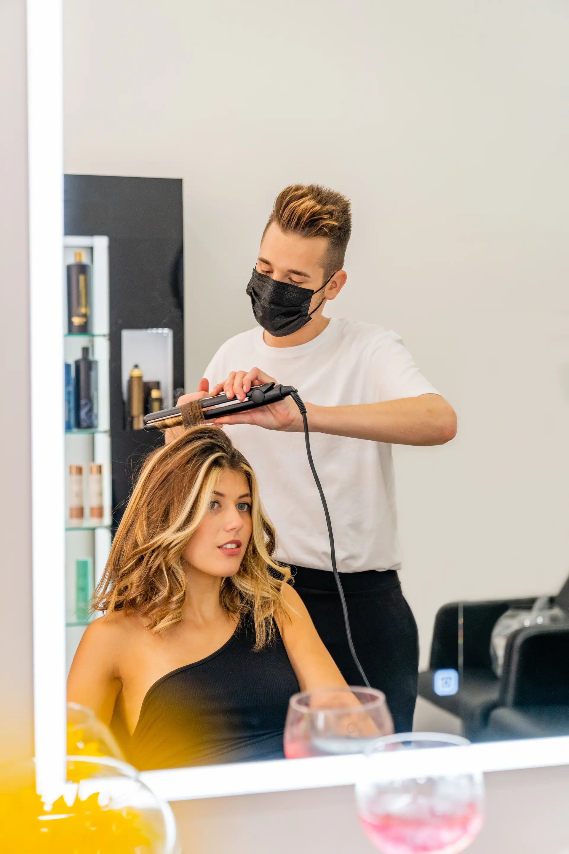 Foto vertical de salón de belleza moderno — interior amplio con hilera de sillas de styling y espejos iluminados, clienta recibiendo color al fondo, maderas claras, plantas y luz natural entrando por ventana grande.